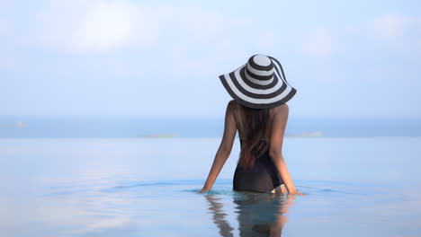 Sexy-woman-with-hat-in-black-One-Piece-swimwear-walking-into-calm-sea