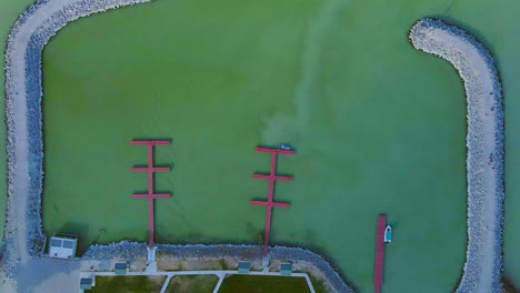 Bird's-eye-view-of-milky-green-water-in-the-Saratoga-Marina-on-Utah-Lake,-two-small-boats-moored-against-red-pontoons-in-the-marina-surrounded-by-a-rocky-wall-with-the-sunlight-catching-the-side