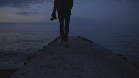 Male-Photographer-Walking-On-The-Concrete-Jetty-By-The-Sea-After-Taking-Photos-On-A-Sunset---ground-level-shot