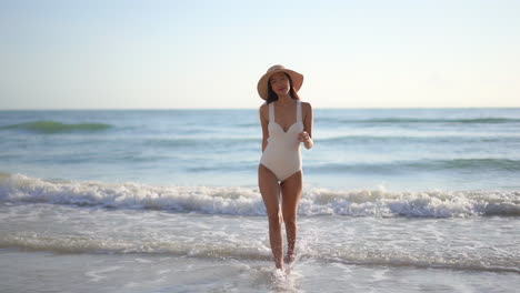Asian-sexy-woman-with-hat-and-in-a-white-one-piece-swimsuit-confidently-comes-out-of-the-ocean-sea-on-the-beach