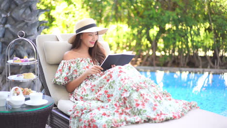 Elegant-young-asian-woman-in-summer-dress-sitting-by-the-pool-reading-the-book