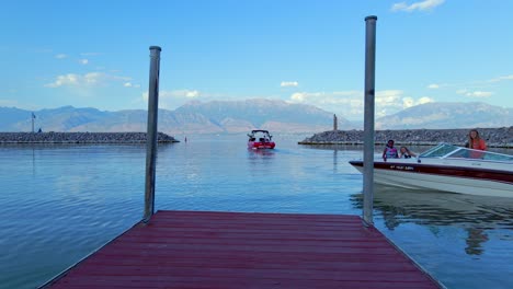 A-family-motorboat-turns-in-front-of-a-wooden-pontoon-ready-to-dock-with-a-bright-red-motorboat-exiting-Saratoga-Springs-marina-in-the-background,-surrounded-by-a-sunlit-mountain-range