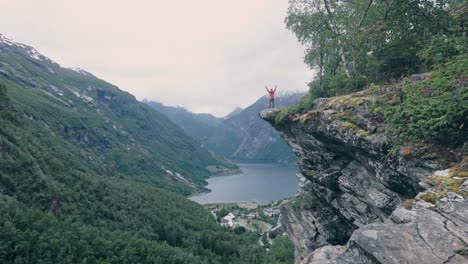 Person-standing-on-the-edge-of-a-mountain,-throwing-arms-up-into-the-air,-surrounded-by-nature,-mountains-and-watching-over-a-beautiful-lake