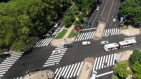 Aerial-of-city-public-transit,-extended-bus-crossing-through-urban-intersection-and-multiple-lanes-of-traffic,-Ben-Franklin-Parkway-in-Philadelphia,-flags-from-around-the-world-along-street
