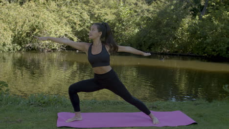 Young-woman-doing-warrior-pose-yoga-exercises-near-pond