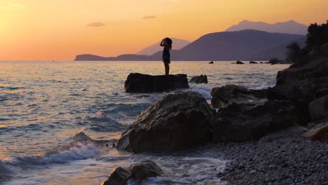 Girl-standing-on-cliff-washed-by-sea-waves-at-beautiful-sunset-with-silhouette-of-mountains-and-orange-sky