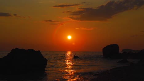 Sonnenuntergangstapete-Mit-Sonne,-Die-Auf-Einem-Farbenfrohen-Himmel-Scheint,-Der-Sich-Durch-Klippensilhouetten-Auf-Lebhaftes-Meerwasser-Widerspiegelt