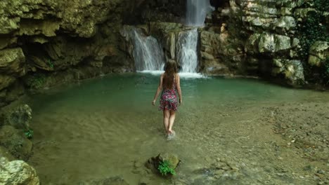 Scenic-landscape-view-above-young-Caucasian-woman-sitting-on-rock-in-shallow-water,-stands-up-and-walks-towards-clear-fresh-Yahshoush-mountain-waterfall,-Lebanon,-behind-overhead-drone-pull-back