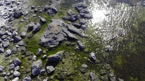 Aerial-view-over-stones-in-shallow-water,-sun-glittering-on-the-water-surface,-on-a-warm,-summer-day,-in-Faro,-Gotland,-Sweden---low,-tilt-up,,-drone-shot
