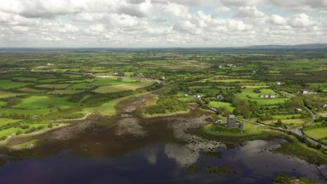 Dunguaire-Castle,-Kinvara,-Galway,-Ireland,-August-2020,-Drone-slowly-pulling-backwards-across-coastal-waters-and-rural-landscape