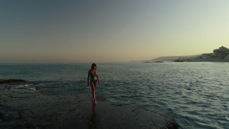 Young-Caucasian-woman-with-long-brown-hair-wearing-bathing-suit-walking-on-Batroun-rock-outcropping-in-blue-ocean-sea-water-at-sunset,-Lebanon,-overhead-aerial-pull-back