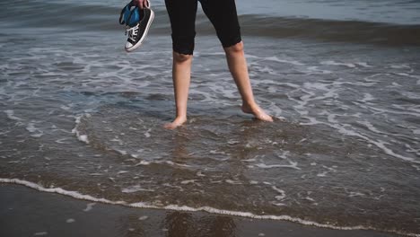 Barefoot-girl-kicking-water-on-beach-holding-sneakers,-slow-motion