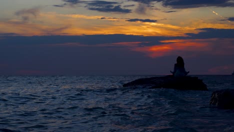 Woman-sitting-on-rock-surrounded-by-dark-sea-water-at-beautiful-sunset,-doing-Yoga-exercises