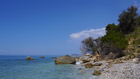 Girl-in-swimsuit-walking-through-rocky-beach-alongside-crystal-water-of-blue-turquoise-sea-in-Greek-island