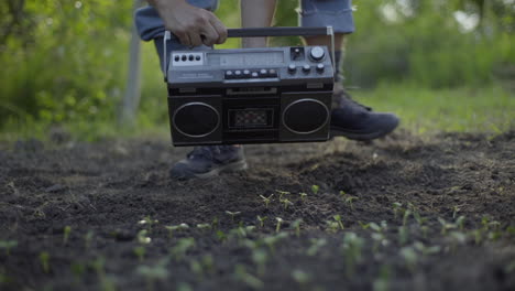 Male-farmer-playing-music-from-vintage-radio-cassette-player-to-green-plants,-backlit-handheld-shot