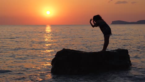 Young-woman-performing-Yoga-concentration-exercises-standing-on-cliff-washed-by-sea-water-at-beautiful-sunset
