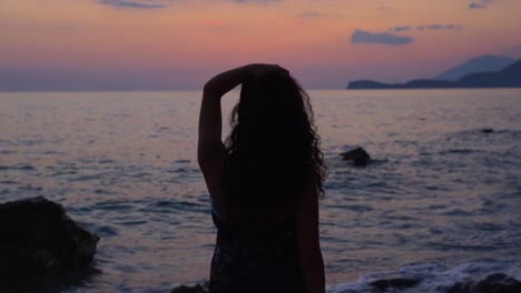 Silhouette-Eines-Mädchens-Mit-Lockigem-Haar,-Das-Am-Felsigen-Strand-Steht-Und-Den-Wunderschönen-Sonnenuntergang-Vor-Dem-Meer-Beobachtet