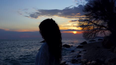 Mädchen-Meditiert-Am-Felsigen-Strand-Und-Beobachtet-Den-Wunderschönen-Sonnenuntergang-Mit-Bewölktem-Himmel,-Der-Sich-Auf-Lebhaftem-Meerwasser-Widerspiegelt
