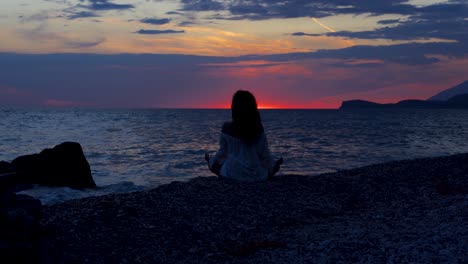 Yoga-pose-of-girl-sitting-on-pebbles-of-tranquil-beach-washed-by-sea-reflecting-colorful-sunset