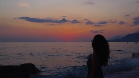 Silhouette-of-beautiful-woman-with-curly-hair-standing-on-rocky-beach,-watching-colorful-sunset