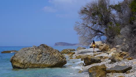 Girl-exploring-tranquil-beach-with-pebbles-and-rocks-splashed-by-sea-waves-in-Ionian-coastline