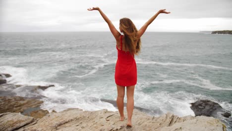 Girl-Raising-Both-Hands-In-The-Sky-While-Enjoying-The-Ocean-Waves-Crashing-On-The-Rocks---Eastern-Suburbs-Beach---Sydney,-NSW,-Australia