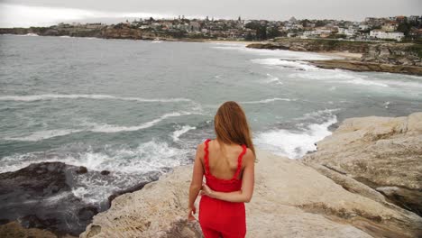 Girl-In-Red-Dress-Walks-Barefoot-On-The-Rocks---Girl-Stands-On-The-Rock-Watches-The-Crashing-Waves---Eastern-Suburbs,-Sydney,-NSW,-Australia