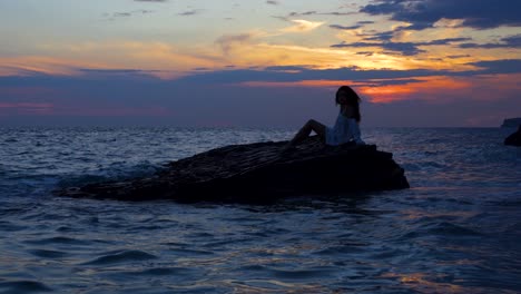 Girl-posing-like-a-siren-over-the-big-cliff-washed-by-sea-and-watching-beautiful-sky-of-the-colorful-sunset