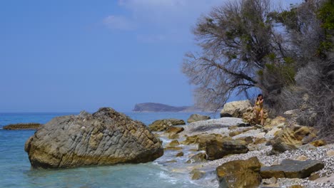 Girl-walking-on-rocky-beach-splashed-by-sea-waves-on-beautiful-seascape-in-Ionian-coastline