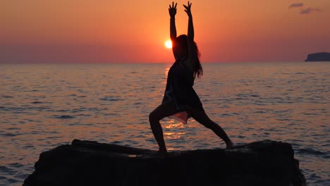 Girl-poses-over-cliff-surrounded-by-vibrant-sea-water-at-beautiful-sunset,-doing-Yoga-exercises