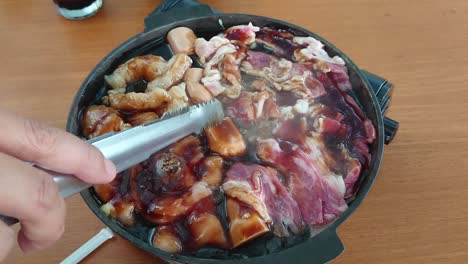 Tasty-delicious-Japanese-style-meats-cooking,-grilling,-sizzling,-and-frying-in-interior-black-pan-with-teriyaki-sauce-in-kitchen,-rotated-with-Caucasian-fingers-holding-silver-tongs,-overhead-static