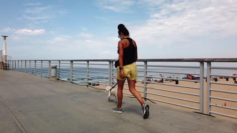 Mujer-Hispana-Caminando-Mascota-De-Perro-Maltés-A-Lo-Largo-Del-Hermoso-Muelle-De-Playa,-California