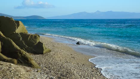 Paradiesstrand-Mit-Kieselsteinen-Und-Felsen,-Die-Von-Wellen-Des-Türkisblauen-Meeres-Umspült-Werden,-Mit-Der-Insel-Korfu-Im-Hintergrund