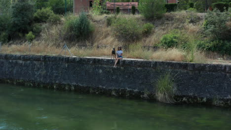 Young-woman-sitting-with-her-dog-along-the-riverbank