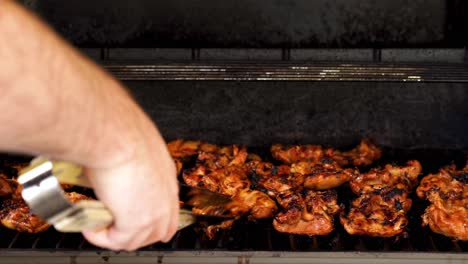 Men's-arm-hand-checking-and-flipping-BBQ-chicken-with-metal-and-wooden-tongs-on-a-gas-grill-as-a-flames-flare-up---still-tripod-shot