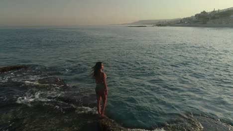 Young-Caucasian-woman-with-long-brown-hair-wearing-bathing-suit-stands-on-Batroun-rock-outcropping-island-in-blue-ocean-sea-water-at-sunset,-Lebanon,-overhead-aerial-flyover-approach