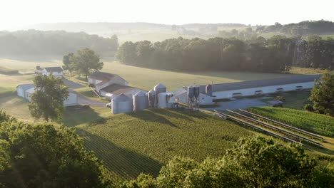 Establishing-shot,-aerial-of-family-farm-in-United-States,-grain-bins,-chicken-house,-barn-and-fields-in-summer-hazy-foggy-morning-light
