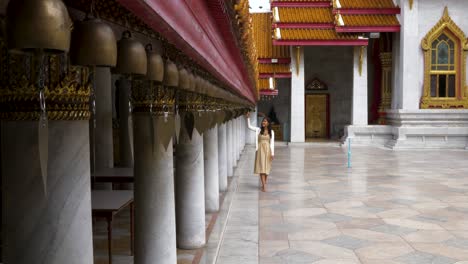 Una-Mujer-Caminando-A-Lo-Largo-De-Las-Campanas-Sagradas-Y-Usando-Su-Mano-Para-Tocarlas-En-Un-Templo-En-Bangkok,-Tailandia