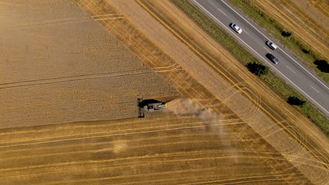 The-harvester-works-in-the-field,-mows-grain-close-to-the-road,-cars-drive-on-the-street,-top-down-aerial-rotate