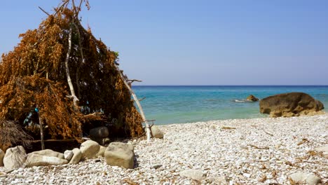 Hut-with-branches-and-leaves-on-secret-beach-with-pebbles-washed-by-clear-crystal-water-of-Ionian-sea