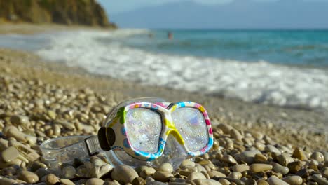 Snorkel-glasses-for-kids-on-pebbles-beach-with-sea-waves-splashing-in-background,-vacation-concept