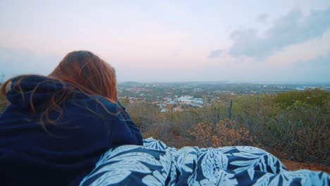 Young-woman-in-hoodie-watching-sunrise-and-laying-on-blanket-at-viewpoint