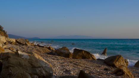Meereswellen,-Die-Auf-Felsen-Eines-Ruhigen-Strandes-Mit-Kiessteinen-Spritzen,-Seelandschaft-Mit-Der-Insel-Korfu-Im-Hintergrund