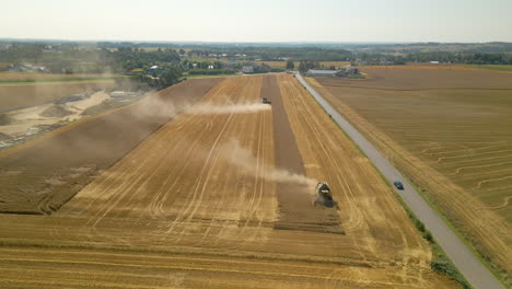 Aerial-backward-flight-showing-harvesting-machines-on-wheat-field-in-middle-of-dust-beside-road-with-driving-car
