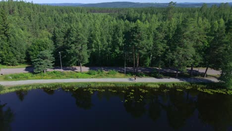 Reversing-drone-footage-over-a-small-lake-showing-two-runners-on-a-gravel-path-with-forest-behind-them