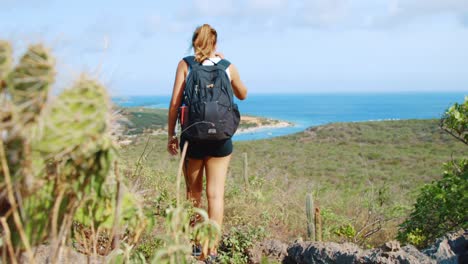 Girl-with-backpack-hiking-down-a-hill-with-coastal-background-in-Curacao,-Caribbean