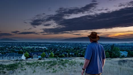 A-man-stands-in-awe-as-a-day-to-night-time-lapse-ZOOMS-OUT-to-reveal-scenes-of-the-distant-mountains-and-a-city-in-a-valley-with-a-lake-in-the-background