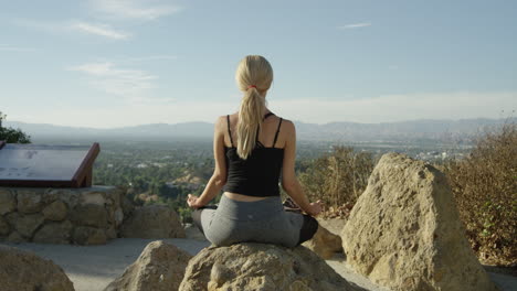 SLOW-MOTION-video-of-young-blonde-woman-seen-from-behind-sitting-on-a-rock-outside-in-yoga-pose-with-blue-sky-and-beautiful-backdrop-views