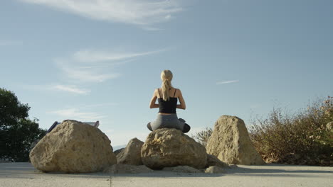 Video-En-Cámara-Lenta-De-Una-Joven-Rubia-Meditando-En-Una-Roca-Afuera-En-Una-Pose-De-Yoga-Con-Cielo-Azul