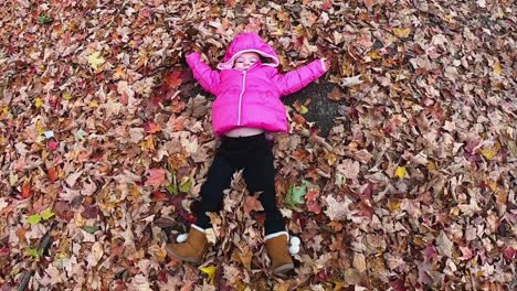 Little-girl-in-pink-hooded-jacket-makes-a-leaf-angel-in-a-carpet-of-fallen-leaves-SLOW-MOTION
